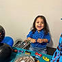 toddler, child, smiling, blue_shirt, toy_cars, cake, chocolate_cake, balloons, party, celebration, table, indoor, happy, curly_hair, person, play, birthday, decorations, jeans, fun