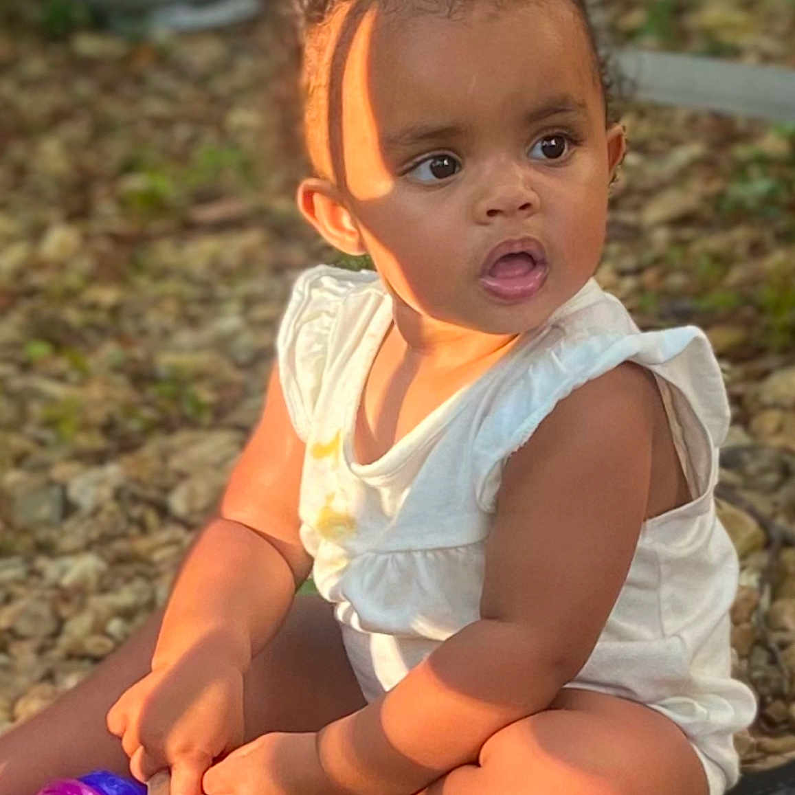 Davina joined the competition — help win amazing prizes! baby, child, curly_hair, expression, face, feet, ground, hands, nature, outdoor, plastic_cup, portrait, rocks, shadow, sitting, skin, sunlight, toddler, toy, white_clothing