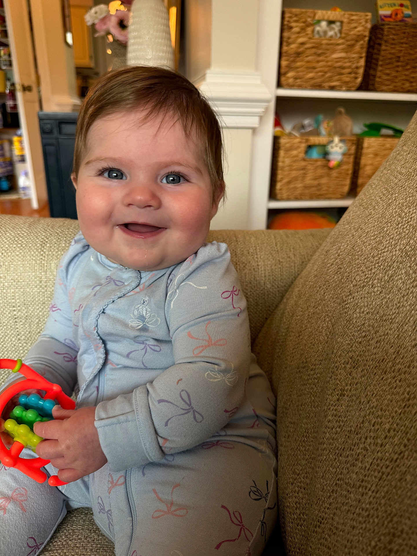 Olivia is registered to the contest to win money with this photo: child, baby, infant, smiling, drooling, toy, onesie, couch, living_room, basket, shelf, blue_eyes, chubby_cheeks, hand, furniture, portrait, cozy, playtime, happy, soft_lighting