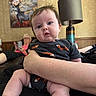 baby, child, person, infant, holding, fox_pattern, clothing, arm, couch, elderly_person, phone, painting, deer, lamp, indoor, living_room, relaxed, seated, curious_expression, home
