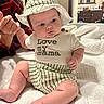 baby, infant, hat, striped_clothing, shirt, hand, adult_hand, plaid_shirt, bed, quilt, indoor, furniture, cabinet, mirror, curious_expression, cute, child, person, family, holding_hand