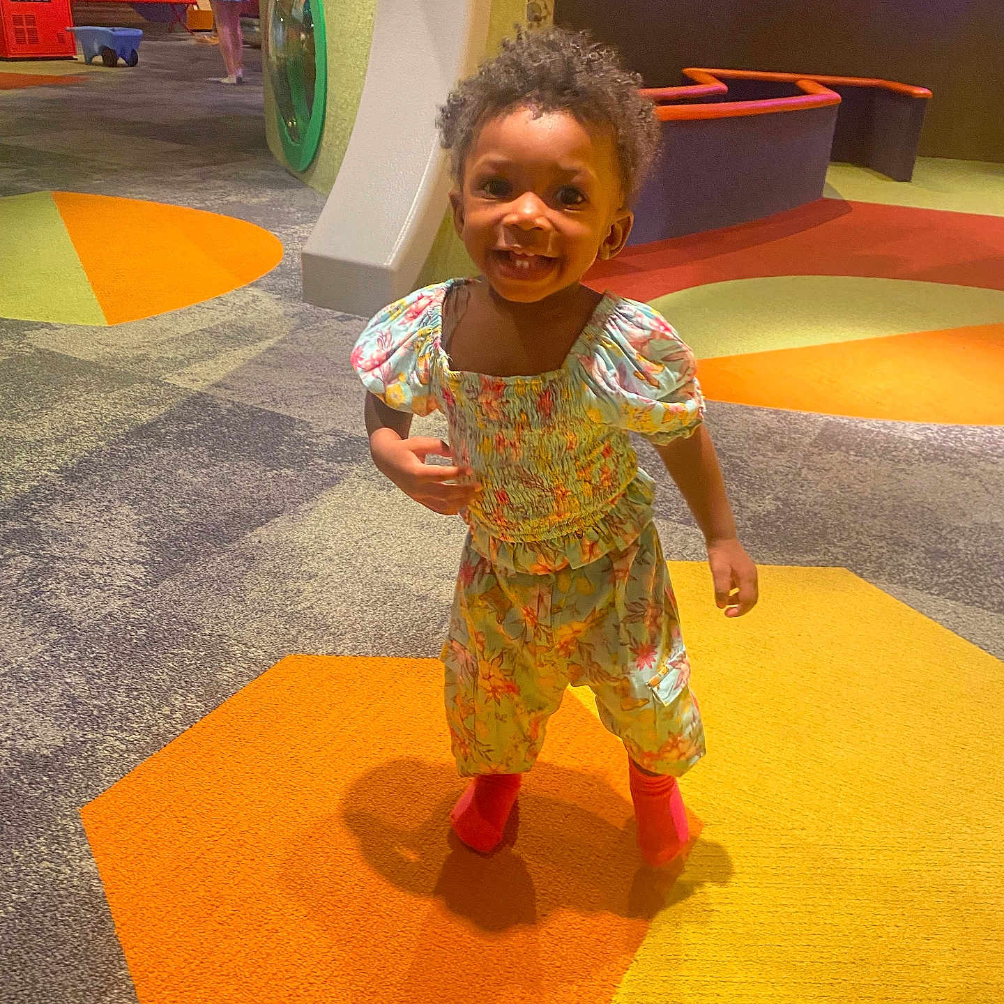 Reign-Diann joined the competition — help win amazing prizes! bright_colors, carpet, child, colorful_carpet, curly_hair, cute, floral_outfit, fun, happy, indoor, orange, patterned_clothing, person, play_area, playground, smiling, standing, toddler, yellow, young_child