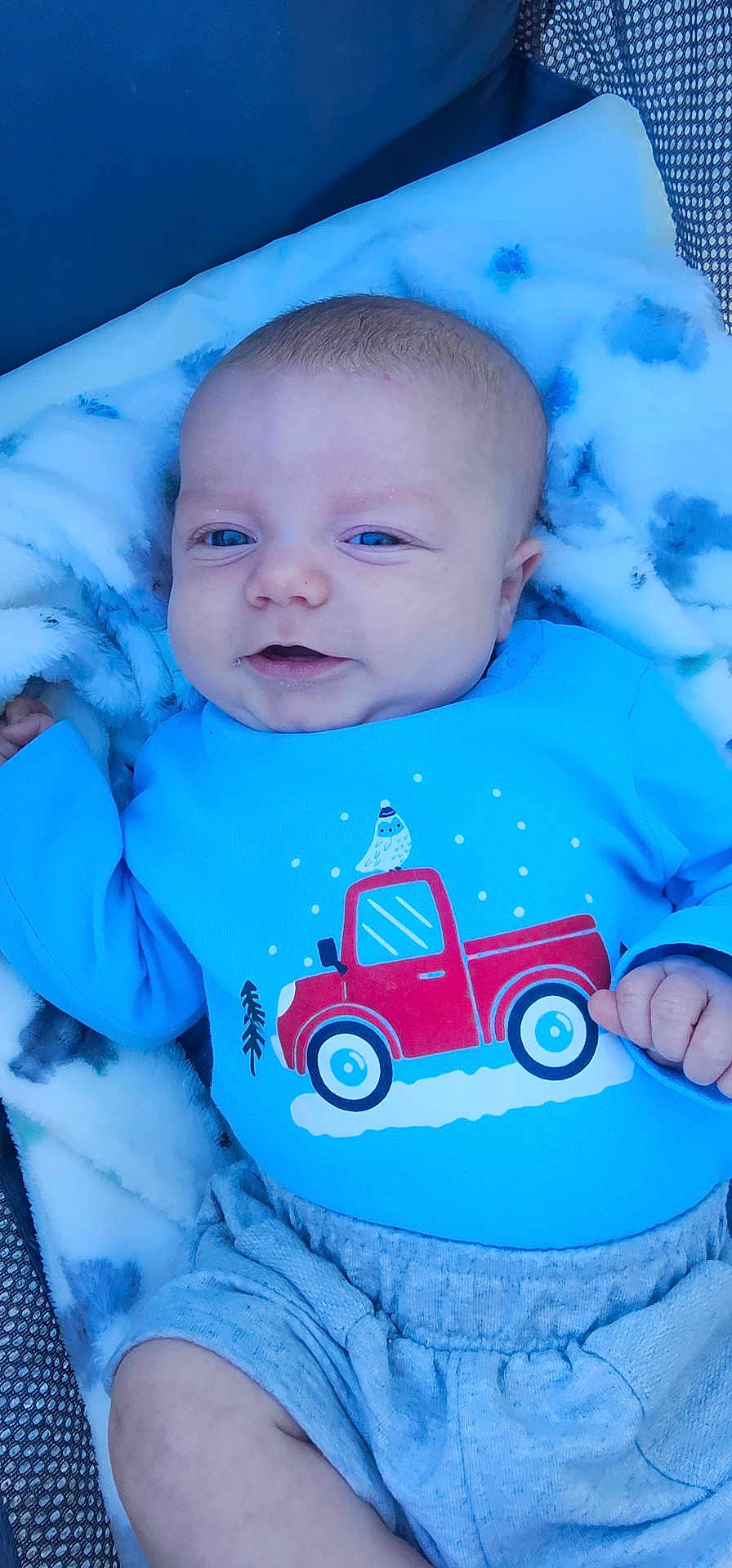 Robbie is registered to the contest to win money with this photo: baby, infant, smiling, blue_shirt, red_truck, blanket, cozy, cute, child, portrait, happy, clothing, lying_down, indoors, soft_texture, young, face, small_hand, casual, comfort