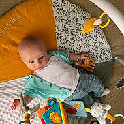 Louka a rejoint le concours — aidez-le/la à gagner de superbes lots ! baby, infant, play_mat, toy, soft_toy, baby_gym, floor, wooden_floor, clothing, jeans, shirt, child, indoor, person, cute, colorful, looking_up, small_toy, pacifier, footwear