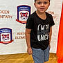 child, boy, portrait, tshirt, shorts, school, background, indoor, smile, face, person, standing, logo, text, shoes, curly_hair, earrings, floor, banner, casual_clothing