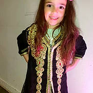 Melya a rejoint le concours — aidez-le/la à gagner de superbes lots ! child, girl, smile, long_hair, hair_accessory, pink_flower, traditional_clothing, embroidered_dress, black_dress, gold_embroidery, standing, hands_on_hips, indoor, wall, confident_pose, person, happy, portrait, fashion, cute