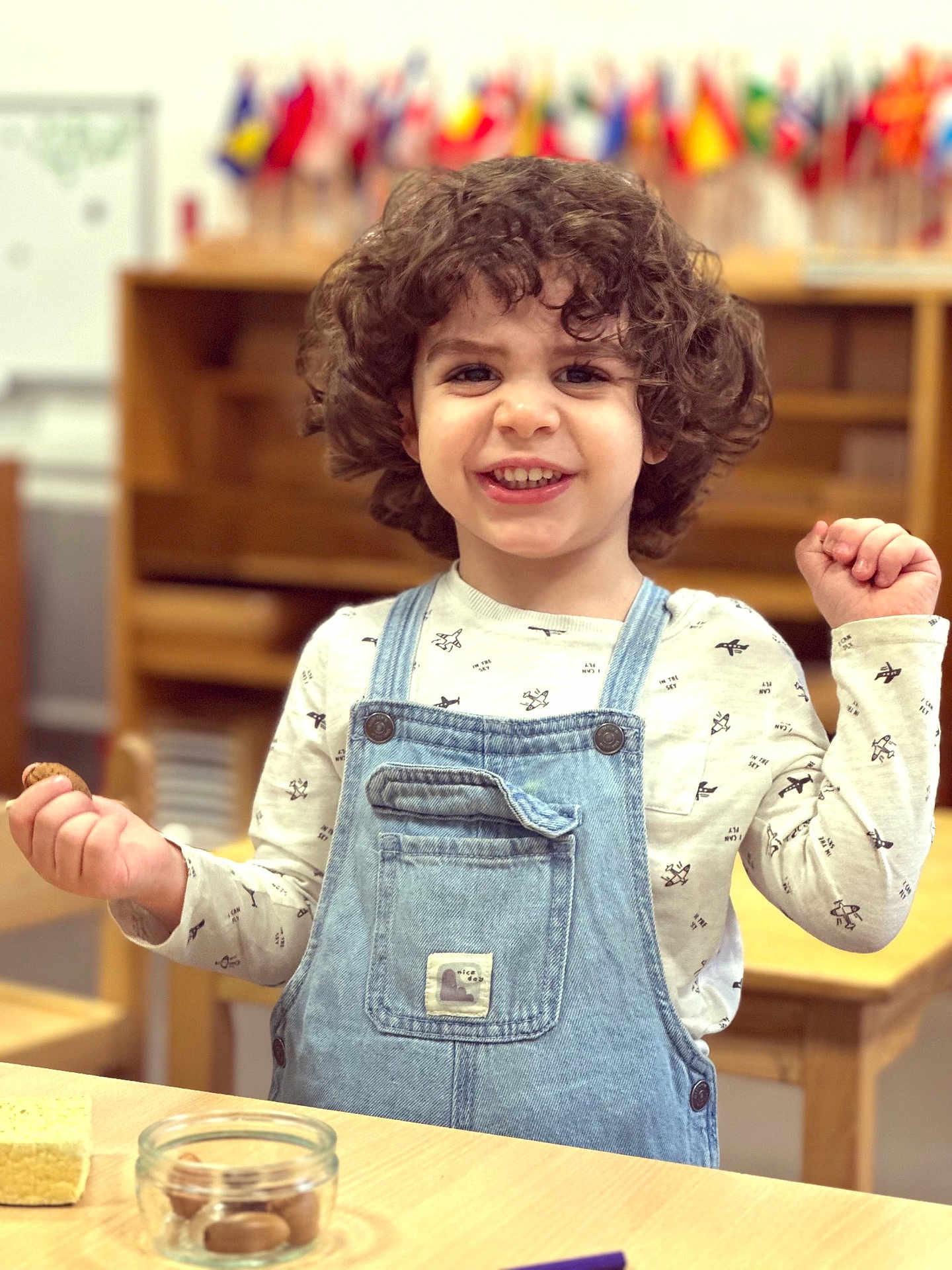 Noa a rejoint le concours — aidez-le/la à gagner de superbes lots ! child, toddler, smiling, curly_hair, denim_overalls, cookie, snack, wooden_table, classroom, flags, happy, indoor, furniture, person, food, long_sleeve_shirt, playful, cute, kid, celebration