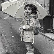 Noa a rejoint le concours — aidez-le/la à gagner de superbes lots ! child, umbrella, raincoat, rain_boots, curly_hair, backpack, sidewalk, street, wet_surface, black_and_white, smiling, urban, rainy_day, dinosaur_boots, portrait, standing, outdoor, person, cute, young