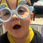 Ilias is registered to the contest to win money with this photo: child, face, glasses, eyewear, indoors, expression, young, person, head, mouth, hair, playful, toy_glasses, furniture, background, bookcase, shirt, clothing, closeup, curious