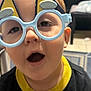 child, face, glasses, eyewear, indoors, expression, young, person, head, mouth, hair, playful, toy_glasses, furniture, background, bookcase, shirt, clothing, closeup, curious