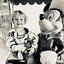 child, toddler, smiling, curly_hair, toy, stuffed_animal, mickey_mouse, statue, black_and_white, indoor, seat, clothing, happy, person, portrait, playful, cute, fun, amusement, kid