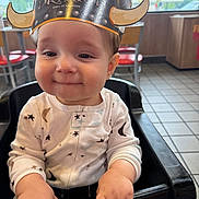 Beau is registered to the contest to win money with this photo: baby, child, high_chair, crown, paper_crown, clothing, onesie, face, hands, indoor, restaurant, table, chair, smile, cute, young_child, playful, portrait, seated, happy