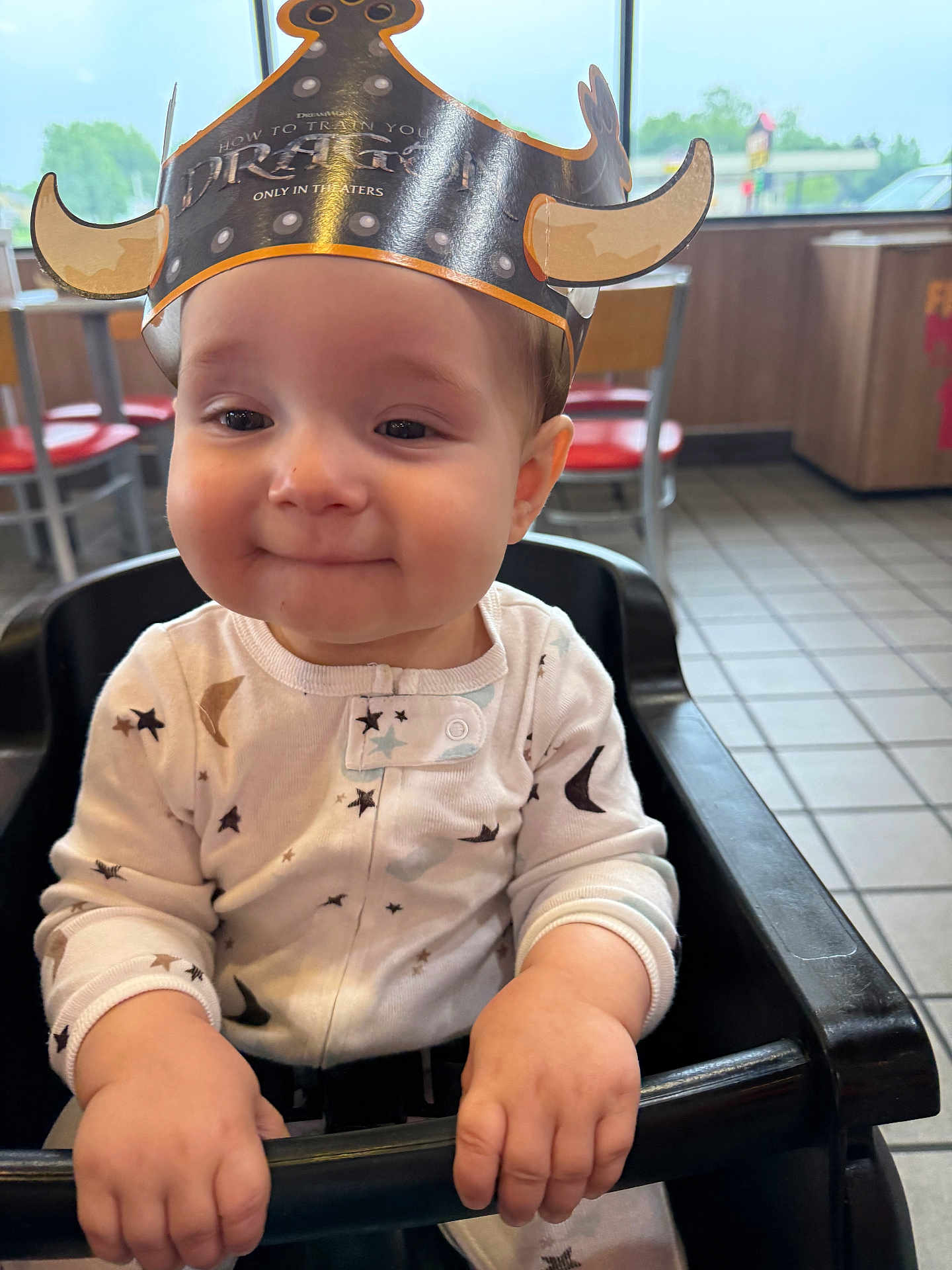 Beau is registered to the contest to win money with this photo: baby, child, high_chair, crown, paper_crown, clothing, onesie, face, hands, indoor, restaurant, table, chair, smile, cute, young_child, playful, portrait, seated, happy