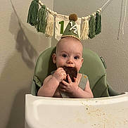 Quentinn joined the competition — help win amazing prizes! baby, high_chair, party_hat, cake, food, messy, celebration, tassel_banner, green_chair, infant, eating, face, hands, indoor, milestone, happy, cute, child, smiling, birthday