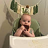 baby, high_chair, party_hat, cake, food, messy, celebration, tassel_banner, green_chair, infant, eating, face, hands, indoor, milestone, happy, cute, child, smiling, birthday