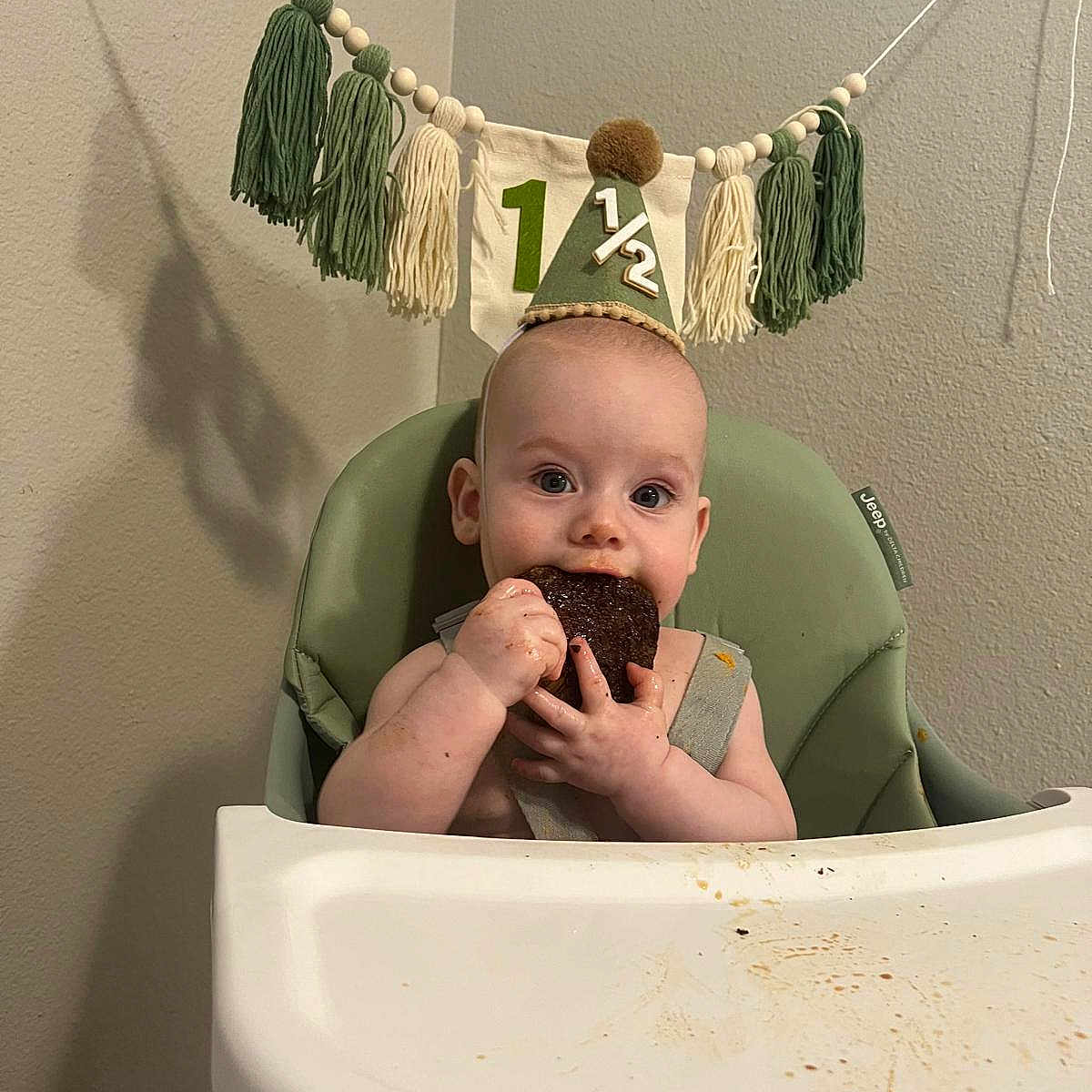 Quentinn joined the competition — help win amazing prizes! baby, high_chair, party_hat, cake, food, messy, celebration, tassel_banner, green_chair, infant, eating, face, hands, indoor, milestone, happy, cute, child, smiling, birthday