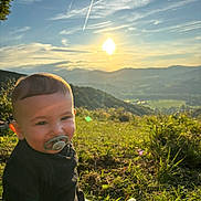 Tino a rejoint le concours — aidez-le/la à gagner de superbes lots ! baby, pacifier, grass, sunset, mountains, outdoor, nature, sky, clouds, sun, hill, child, greenery, landscape, smiling, casual_clothing, daylight, scenic, portrait, relaxation