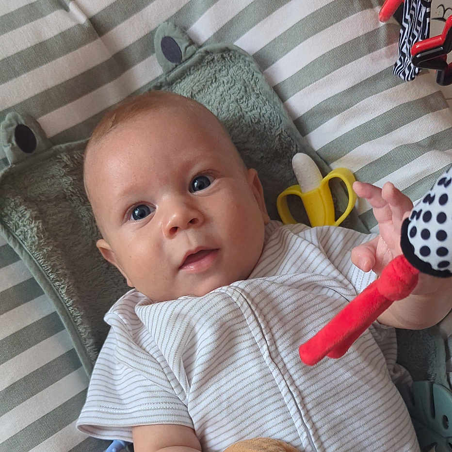 Zaiyan is registered to the contest to win money with this photo: baby, banana_toy, blue_eyes, child, close_up, comfort, face, frog_pillow, hand, indoor, infant, lying_down, person, plush_pillow, polka_dot, red_toy, short_sleeves, striped_cushion, toy, white_clothing