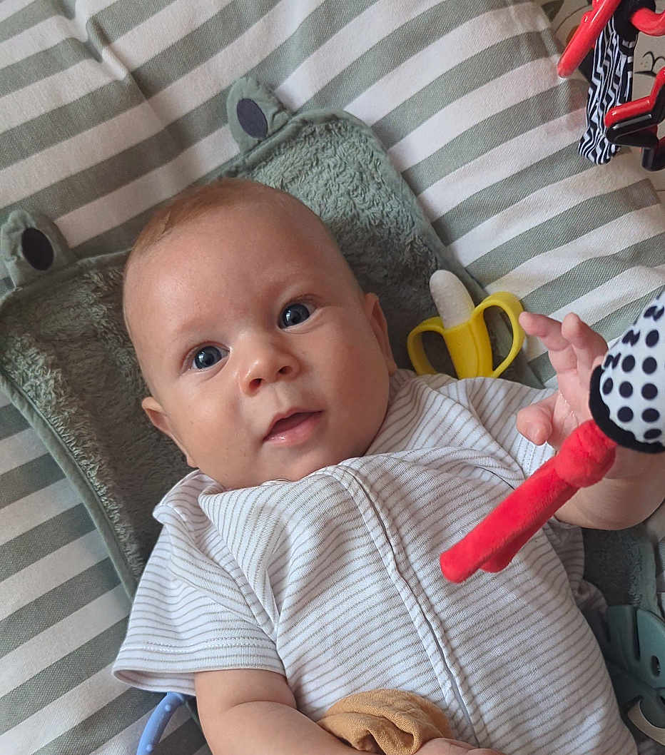 Zaiyan is registered to the contest to win money with this photo: baby, infant, blue_eyes, striped_cushion, plush_pillow, frog_pillow, toy, banana_toy, polka_dot, red_toy, white_clothing, short_sleeves, face, hand, lying_down, indoor, close_up, child, person, comfort