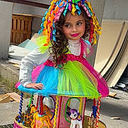 Ancana a rejoint le concours — aidez-le/la à gagner de superbes lots ! child, girl, colorful_costume, tutu, carousel, toy_horse, curly_hair, party_hat, makeup, bright_colors, playful, portrait, outdoor, concrete, construction_materials, pink, green, blue, smile, festive