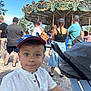 Sacha participe au concours pour gagner de l'argent avec cette photo : child, boy, cap, white_shirt, carousel, outdoor, daylight, people, crowd, bench, stroller, pavement, trees, summer, smile, face, portrait, park, casual_clothing, funfair