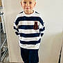 child, boy, smiling, striped_sweater, navy_pants, socks, indoor, wooden_floor, wall, casual_clothing, hands_behind_back, cute, portrait, standing, short_hair, light_skin, happy, home, flooring, playful