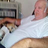 elderly_man, dog, indoor, chair, relaxing, white_shirt, pet, home, furniture, human, animal, resting, blurred, hand, clock, remote_control, casual, seated, living_room, comfort