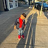 child, boy, smiling, outdoor, sidewalk, street, urban, portrait, standing, fashion, jacket, red_shirt, jeans, sneakers, red_shoes, parked_cars, storefronts, trees, shadows, dog_waste_sign