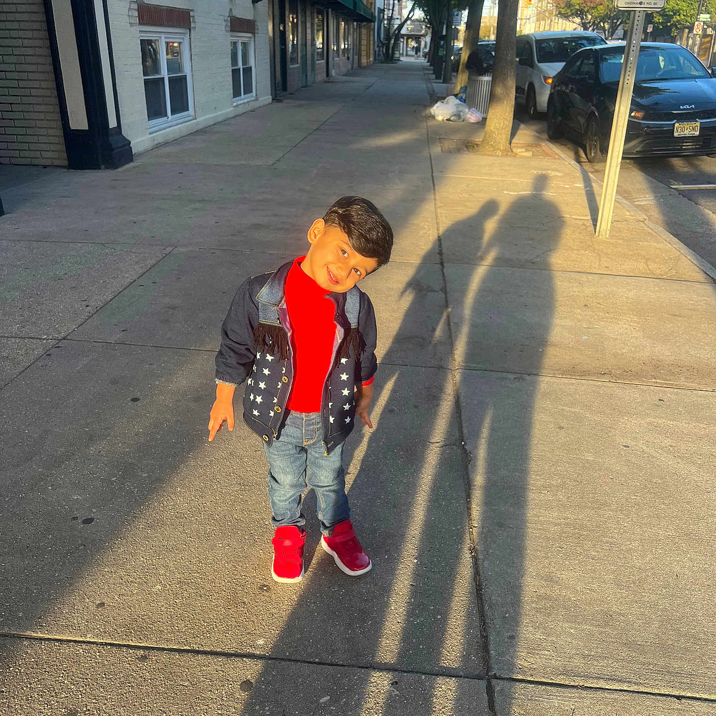 Krishiv is registered to the contest to win money with this photo: child, boy, smiling, outdoor, sidewalk, street, urban, portrait, standing, fashion, jacket, red_shirt, jeans, sneakers, red_shoes, parked_cars, storefronts, trees, shadows, dog_waste_sign