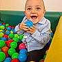 baby, child, ball_pit, blue_ball, colorful_balls, happy, smiling, blue_eyes, light_blue_shirt, dark_pants, indoor, playtime, cute, small_hands, joyful, seated, toy, fun, infant, playground