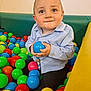 Riley is registered to the contest to win money with this photo: toddler, child, ball_pit, blue_ball, colorful, playful, happy, indoor, toy, person, clothing, smile, face, seated, playtime, cute, bright_colors, young_child, casual_clothing, fun