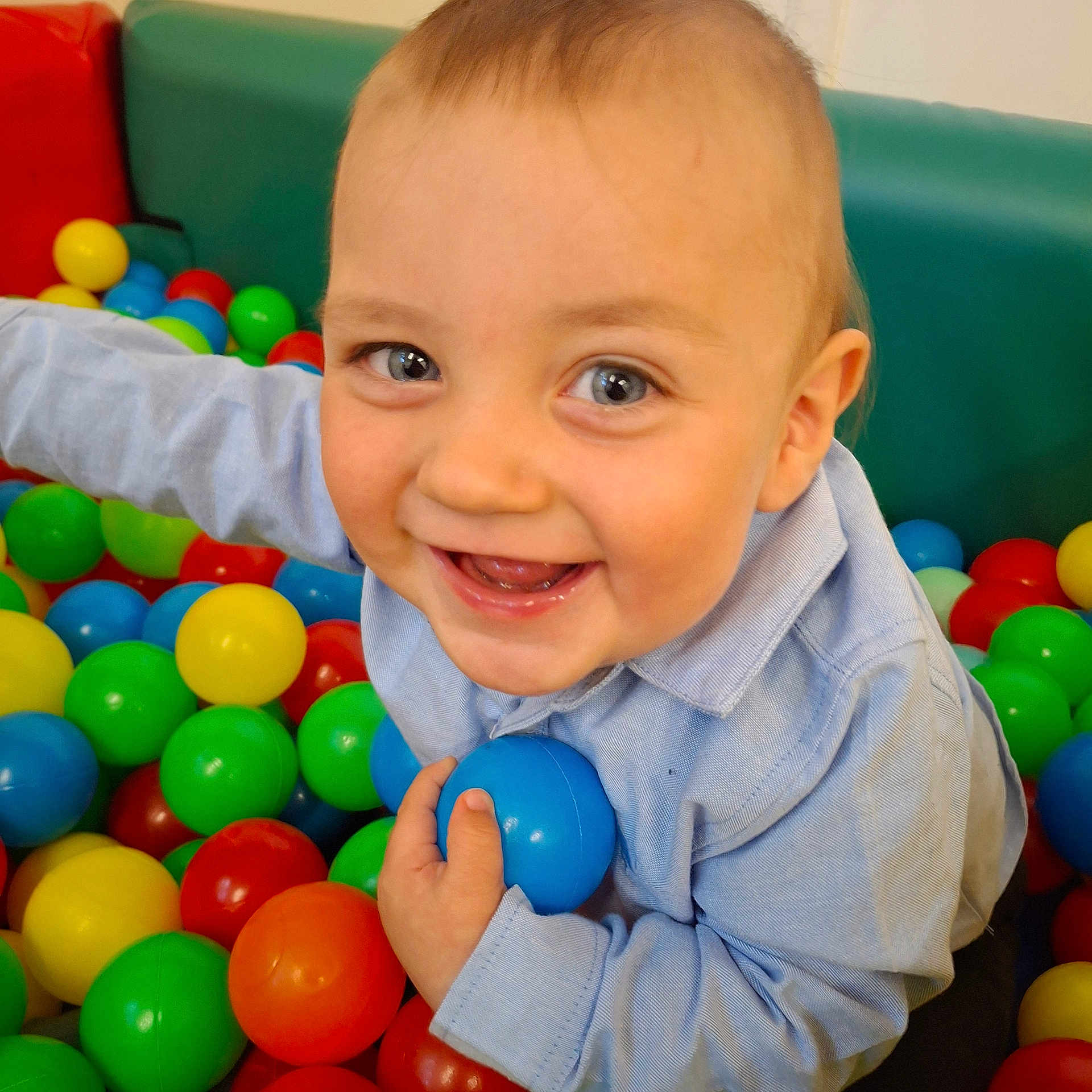 Riley joined the competition — help win amazing prizes! baby, child, toddler, smiling, happy, blue_ball, colorful_balls, play, indoor, casual_clothing, light_blue_shirt, face, eyes, hand, fun, joy, cute, background, green_cushion, red_cushion