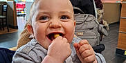Riley is registered to the contest to win money with this photo: baby, child, smiling, eating, indoor, restaurant, high_chair, clothing, face, hand, food, table, bag, stroller, happy, person, apparel, sitting, infant, casual