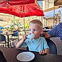 child, boy, blond, espresso, cup, cafe, outdoor, table, chair, tattoo, red_umbrella, daylight, clothing, person, drink, plate, casual, curious, blue_eyes, short_sleeves