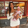 woman, baby, child, holding, white_dress, pink_headband, pink_socks, outdoor, window, red_frame, reflection, portrait, person, smile, curly_hair, casual, daylight, concrete_floor, wooden_chair, basket