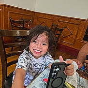 Ethan is registered to the contest to win money with this photo: child, smiling, phone, smartphone, table, wooden_chair, indoor, happy, toddler, casual_clothing, person, face, hair, hand, furniture, plaid_tablecloth, dining_area, young_child, interior, playful