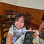 child, smiling, phone, smartphone, table, wooden_chair, indoor, happy, toddler, casual_clothing, person, face, hair, hand, furniture, plaid_tablecloth, dining_area, young_child, interior, playful