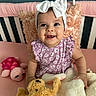 baby, child, smiling, headband, bow, plush_toy, teddy_bear, bunny, bed, pillow, pink, purple, dress, infant, cute, happy, indoors, toy, person, portrait