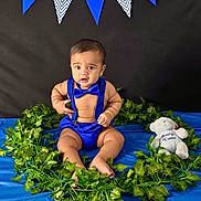 Shaurya is registered to the contest to win money with this photo: baby, banner, black_background, blue_background, blue_clothing, bow_tie, child, cute, decorations, expression, fabric, green_leaves, indoor, infant, plush_toy, portrait, sitting, suspenders, teddy_bear, wreath