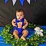 baby, banner, black_background, blue_background, blue_clothing, bow_tie, child, cute, decorations, expression, fabric, green_leaves, indoor, infant, plush_toy, portrait, sitting, suspenders, teddy_bear, wreath