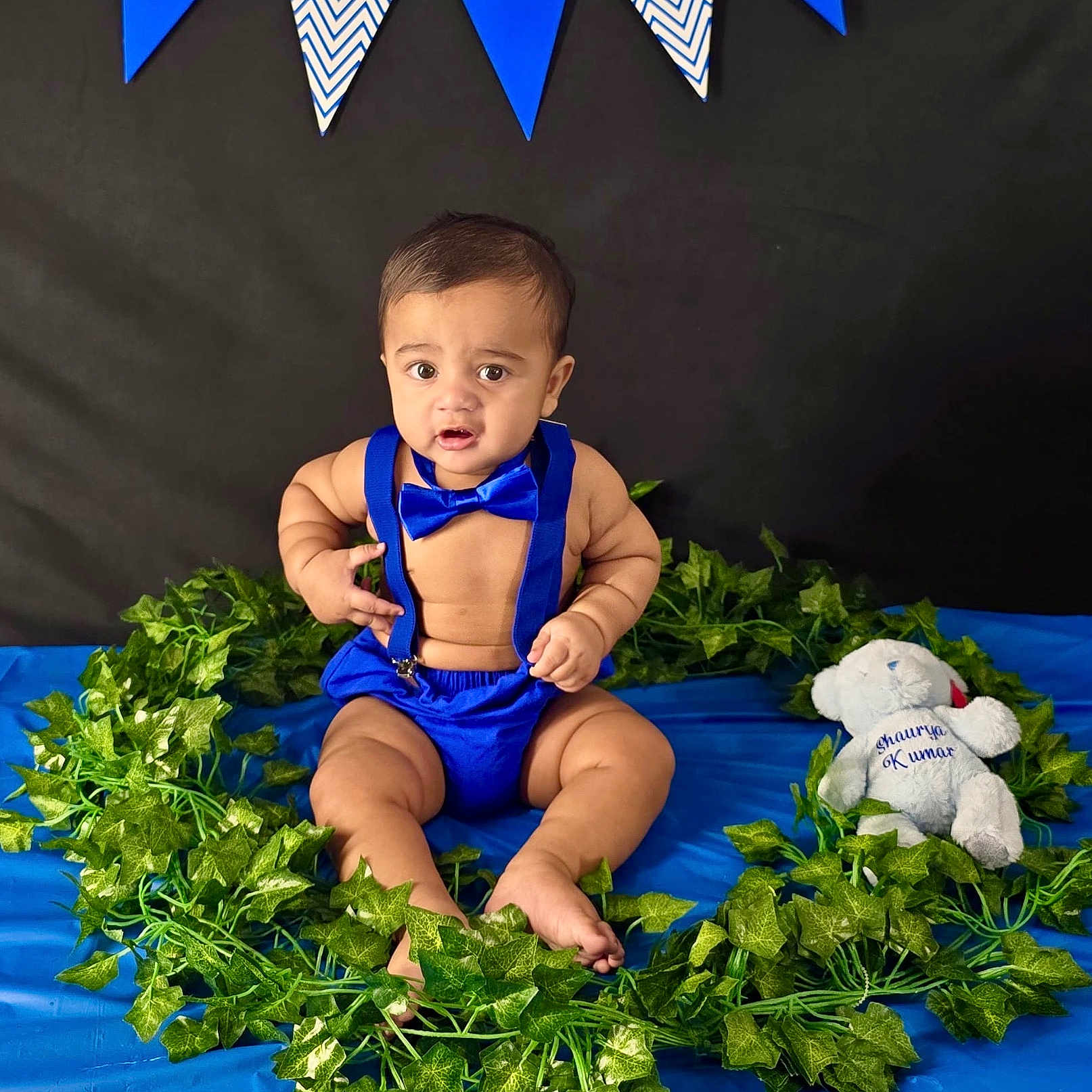 Shaurya is registered to the contest to win money with this photo: baby, banner, black_background, blue_background, blue_clothing, bow_tie, child, cute, decorations, expression, fabric, green_leaves, indoor, infant, plush_toy, portrait, sitting, suspenders, teddy_bear, wreath