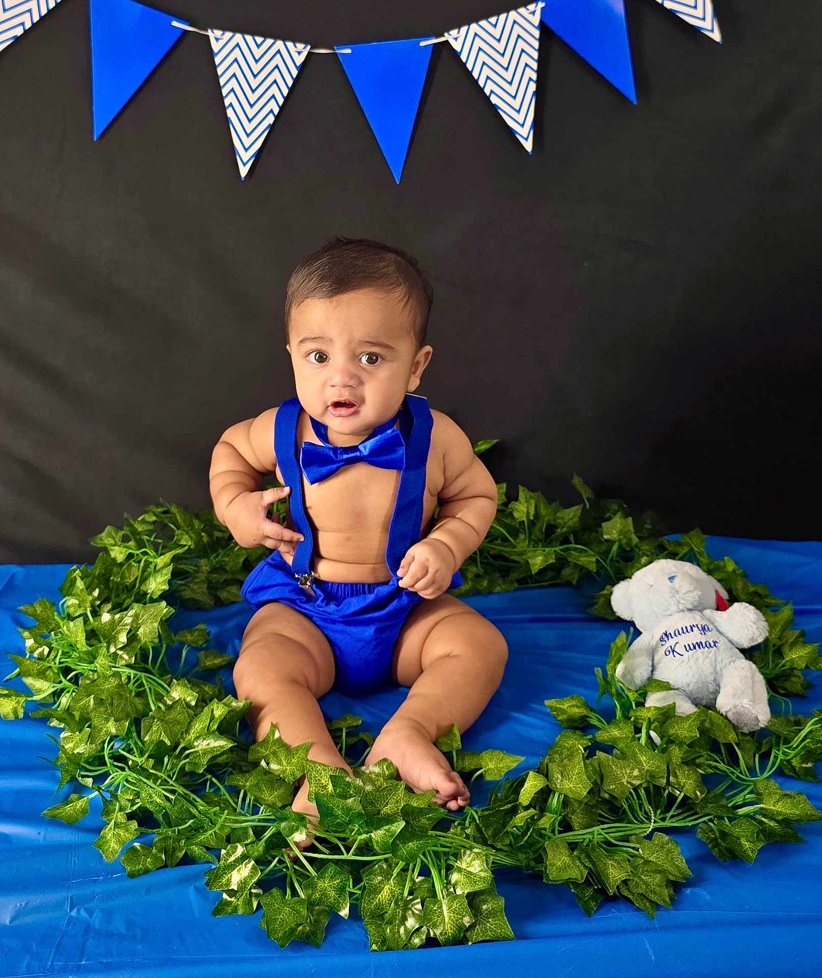 Shaurya is registered to the contest to win money with this photo: baby, banner, black_background, blue_background, blue_clothing, bow_tie, child, cute, decorations, expression, fabric, green_leaves, indoor, infant, plush_toy, portrait, sitting, suspenders, teddy_bear, wreath
