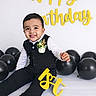 1st_birthday, baby, birthday, black_balloons, celebration, child, cute, decorations, flower_corsage, formal_wear, happy_birthday, indoors, joyful, party, portrait, sitting, smiling, studio, suit, yellow_letters