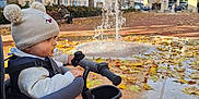Leo a rejoint le concours — aidez-le/la à gagner de superbes lots ! toddler, child, tricycle, pacifier, hat, pom_poms, autumn, leaves, fountain, park, bench, elderly_person, sunny, outdoor, building, sidewalk, jacket, shoe, person, daytime