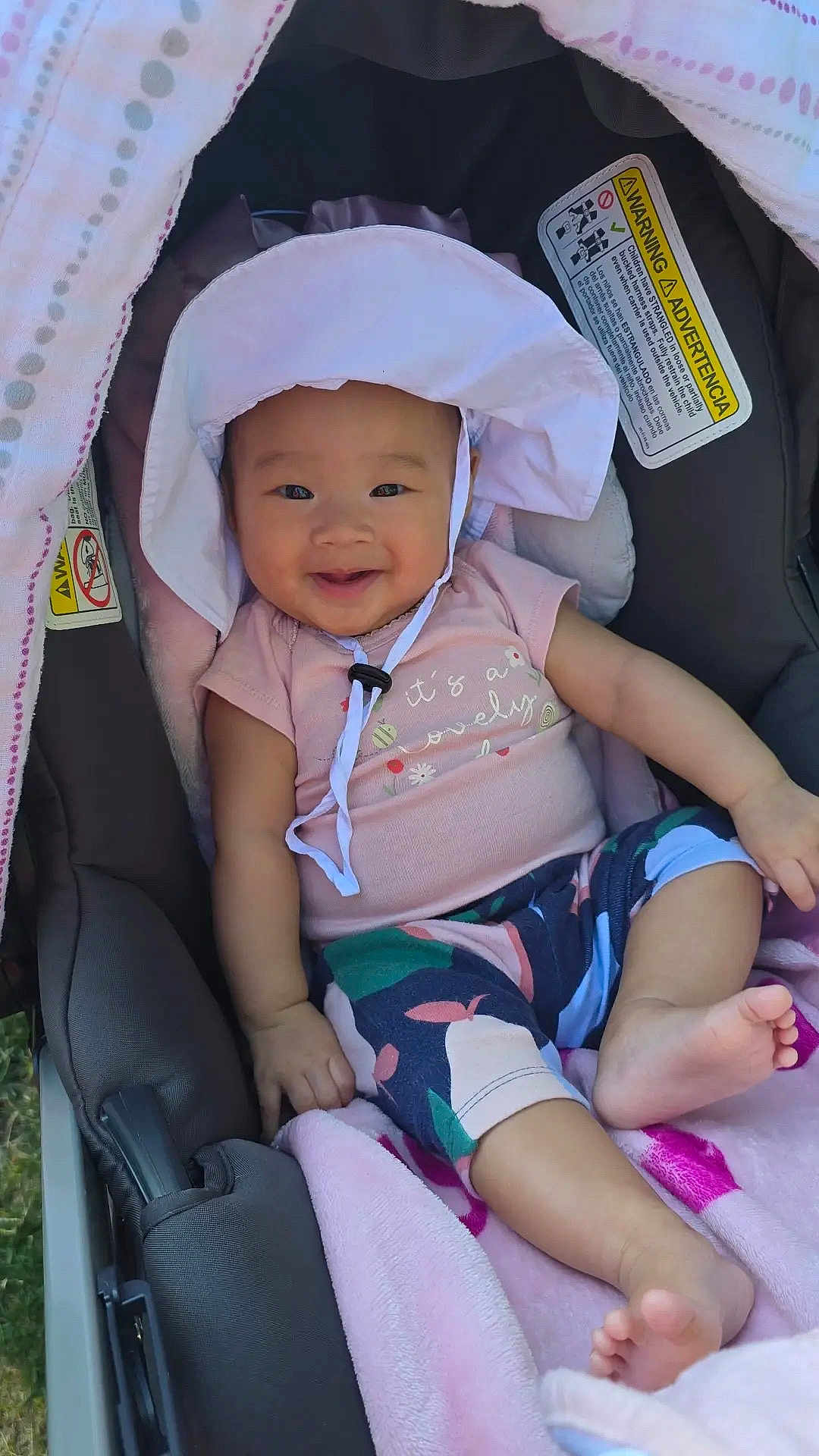 Everly is registered to the contest to win money with this photo: baby, child, smiling, hat, stroller, blanket, clothing, infant, cute, outdoor, person, face, happy, legs, feet, sunhat, patterned_pants, seat, comfort, relaxing