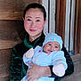 Devick is registered to the contest to win money with this photo: woman, baby, smile, holding, green_velvet, hat, mittens, window, wooden_wall, indoor, portrait, person, clothing, face, happy, family, warm_clothes, infant, cute, mother