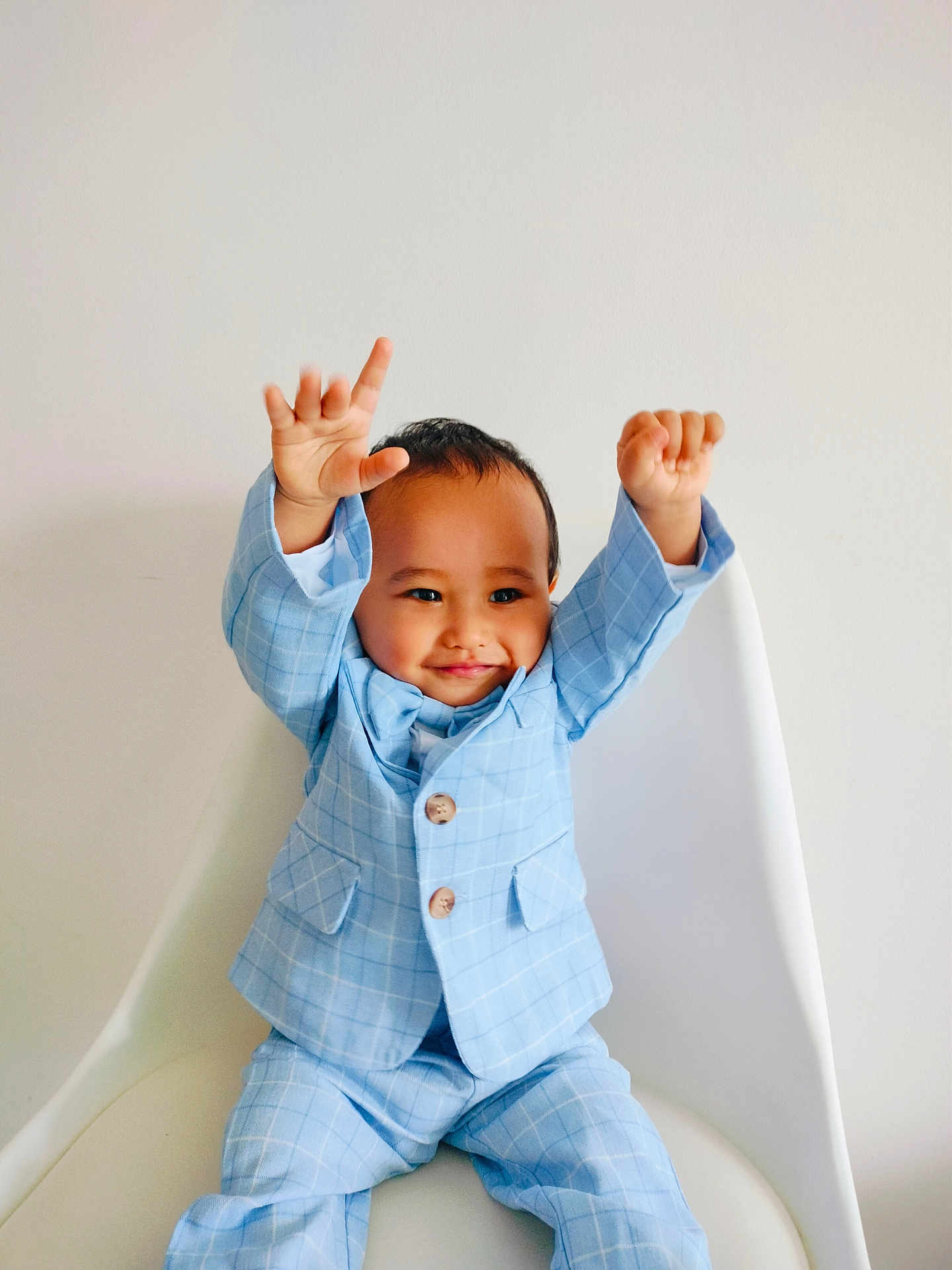 Devick joined the competition — help win amazing prizes! toddler, child, smiling, blue_suit, checkered_suit, raised_hands, white_chair, indoor, portrait, happy, cute, fashion, casual, person, young_child, baby_face, seated, celebration, joy, minimalist_background