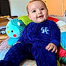baby, blue_onesie, child, cushion, stuffed_animal, toy, indoor, smiling, happy, person, face, head, seated, plush, colorful, home, furniture, door, wall_decor, cozy