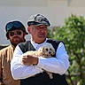 animal, beard, casual, daylight, dog, flat_cap, friendly, greenery, helmet, holding, human, man, nature, outdoor, pet, portrait, sunglasses, two_people, vest, white_dog
