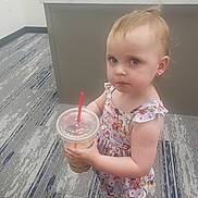 Mila is registered to the contest to win money with this photo: toddler, child, drink, straw, dress, princess, earrings, carpet, office, desk, floor, footwear, indoor, person, young_child, serious_expression, holding, beverage, plastic_cup, standing